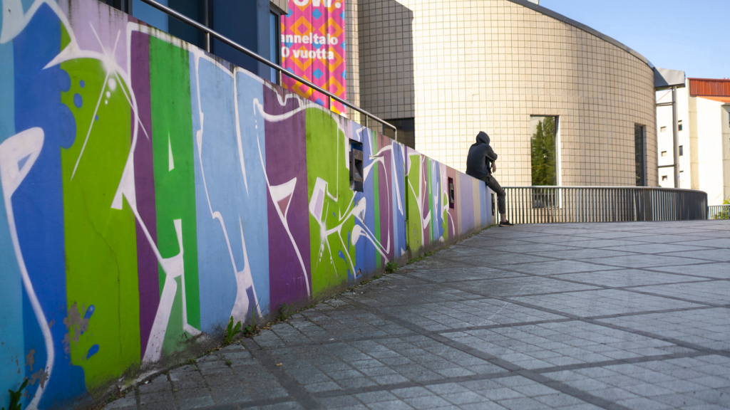 Huppuipäinen nuori istuu graffitoidulla muurilla selkä kameraa kohti.